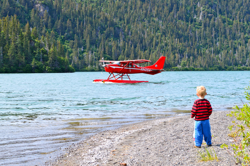 A toddler standing on the beach, looking at a red floatplane slowly motoring by on Leisure Lake. 